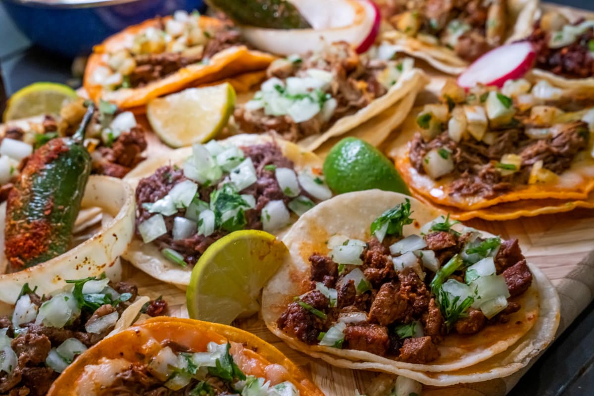 Assortiment de délicieux tacos authentiques, bière, carne asada, adobada, cabeza et chicharone, disposés avec des tranches de citron vert, de l'oignon et du piment rôti